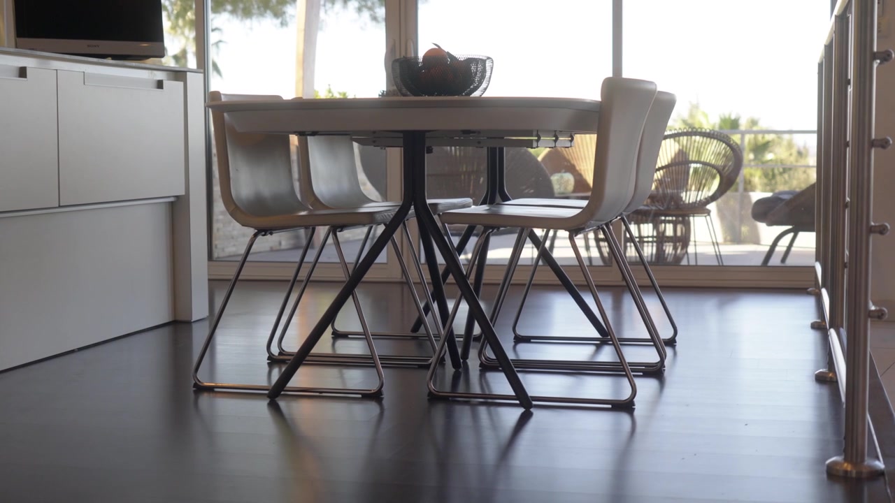 Langsomt bueskud af et moderne rundt spisebord med stole med metalben på mørkt trægulv inde i et moderne køkken og et stort vindue. Klart dagslys gennem gulv-til-loft-vinduer.