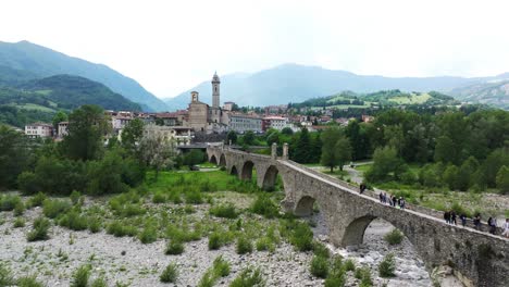 People-crossing-Ponte-Gobbo-with-Bobbio-small-town-and-mountains-in-background,-Piacenza-province-of-Emilia-Romagna-in-northern-Italy