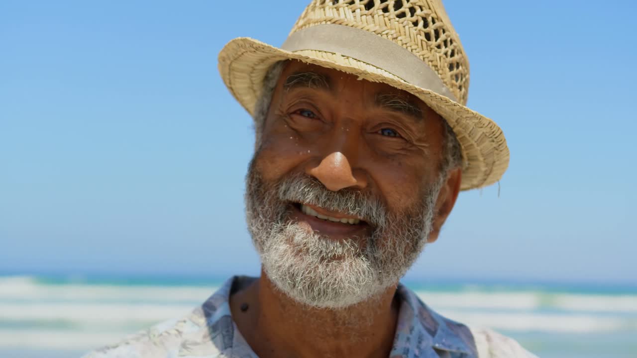 vista frontal de un hombre afroamericano sénior activo con sombrero de pie en la playa al sol 4k