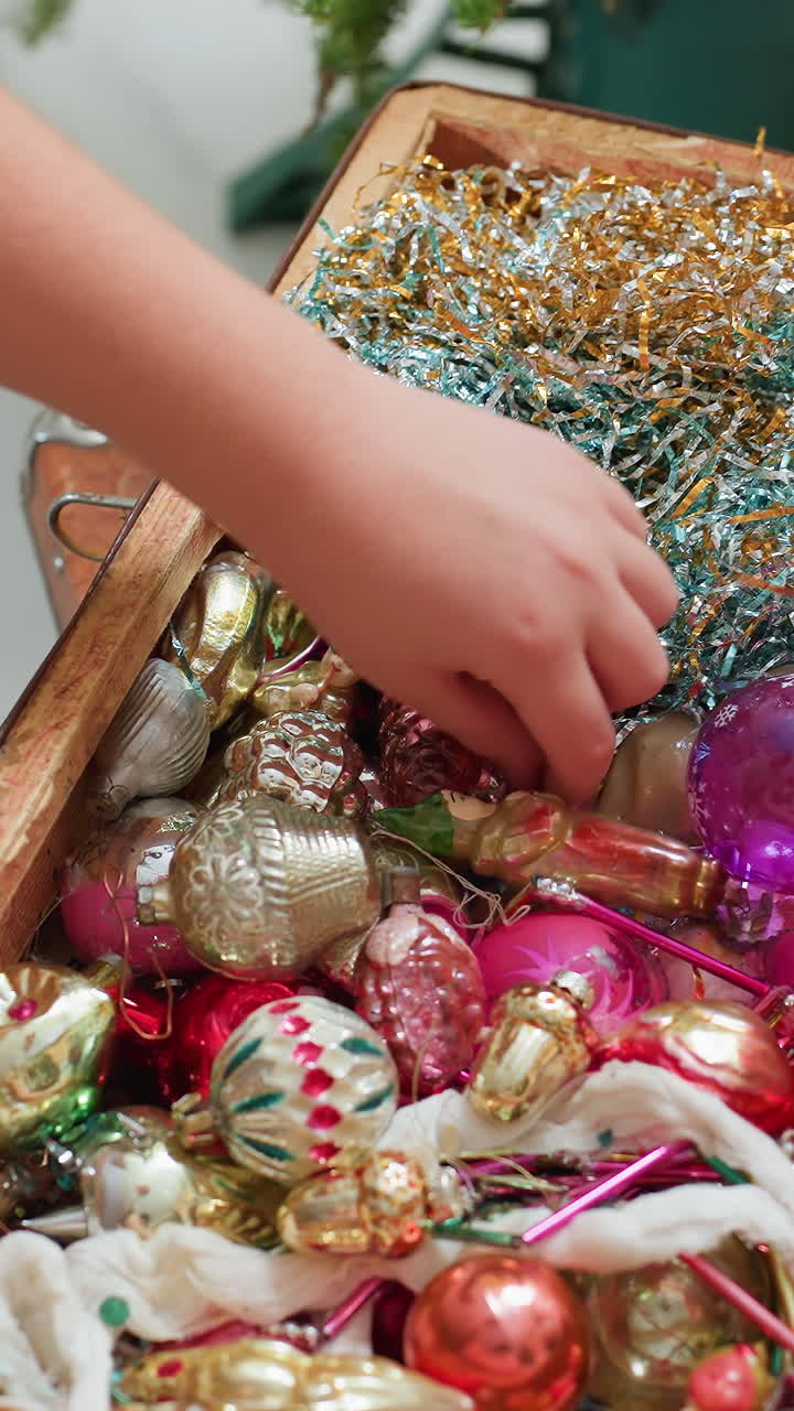 Un niño abre una caja de adornos vintage, unas pequeñas manos rebuscan entre espumillón y bolas de cristal, decoraciones en rosa pastel y dorado, borde del baúl cubierto de polvo, un acogedor salón y manipulación suave y cuidadosa.