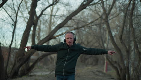 a man in a green jacket and headphones stretches his arms wide in a forest, dancing to music he is listening to