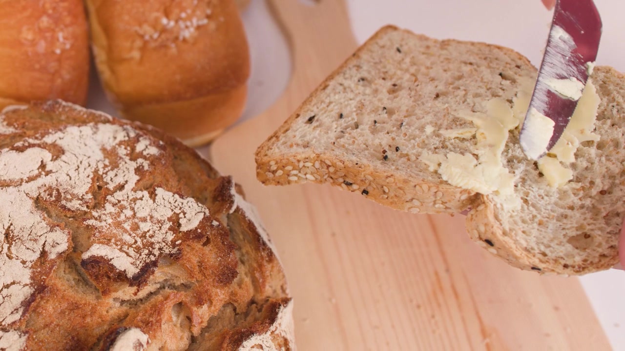 Primo piano di un coltello che spalma burro su pane ai cereali tra diverse pagnotte artigianali.