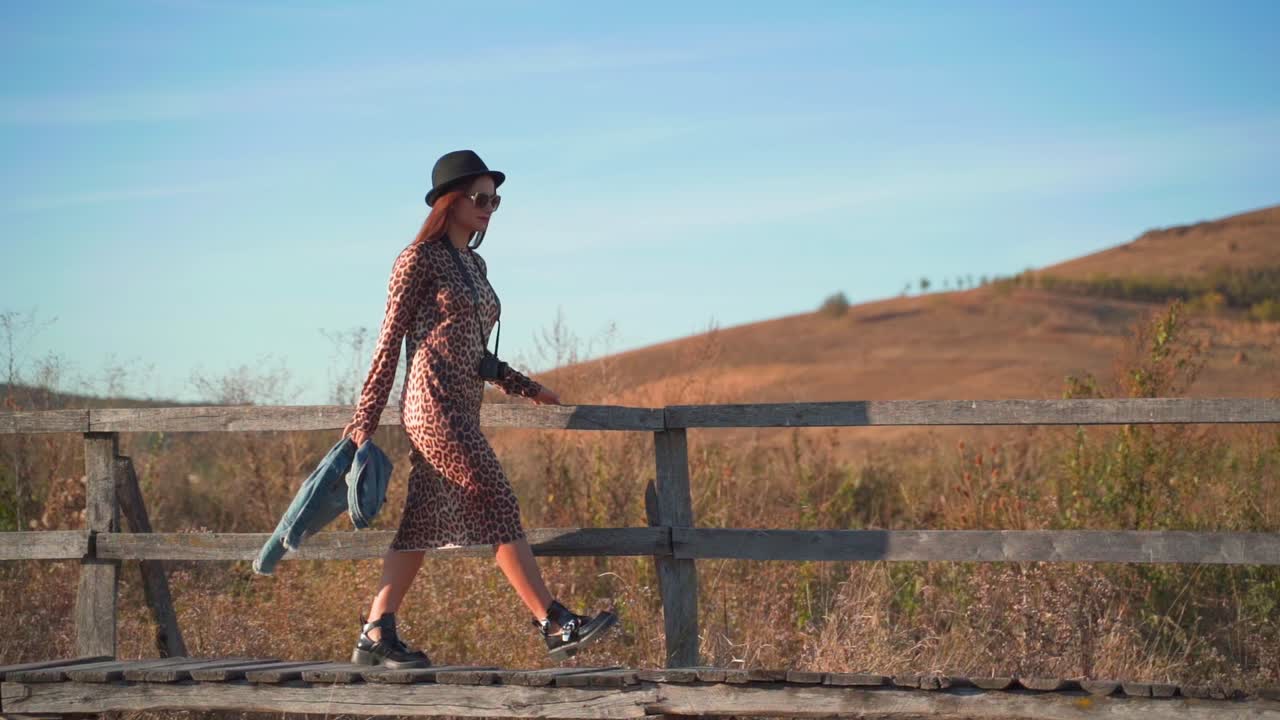 Young Caucasian woman wearing black hat and fashionable printed animal dress carrying jean jacket walks on wooden boardwalk, handheld slow motion
