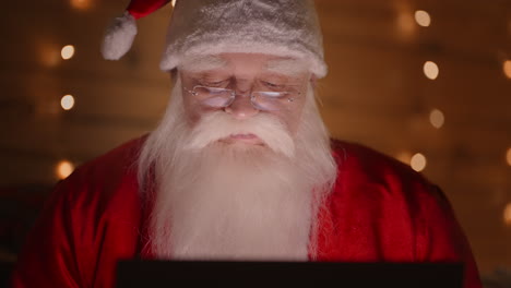 front view a real santa claus is working with a laptop at night with glasses in the light of christmas lights on the background of a christmas tree. remote work