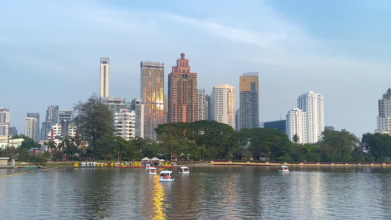 Paddle boats glide across a serene lake in Benjakitti Park, Bangkok, with a city skyline backdrop under soft daylight