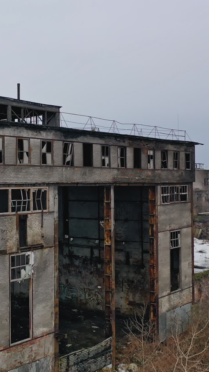 Old abandoned plant with broken windows ruined after the military actions. Demolished plant with pitted roof and some snow on the ground in winter. Aerial view Vertical video
