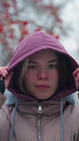 primer plano de una joven ajustando su gorra con capucha mientras mira directamente a la cámara con expresión seria, de pie al aire libre en chaqueta de invierno, árboles borrosos en el fondo