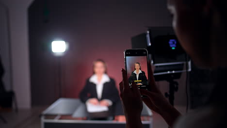 beautiful caucasian lady with long hair wearing a white shirt and black jacket on the phone screen. female reporter is getting ready for footage in studio. blurred backdrop.