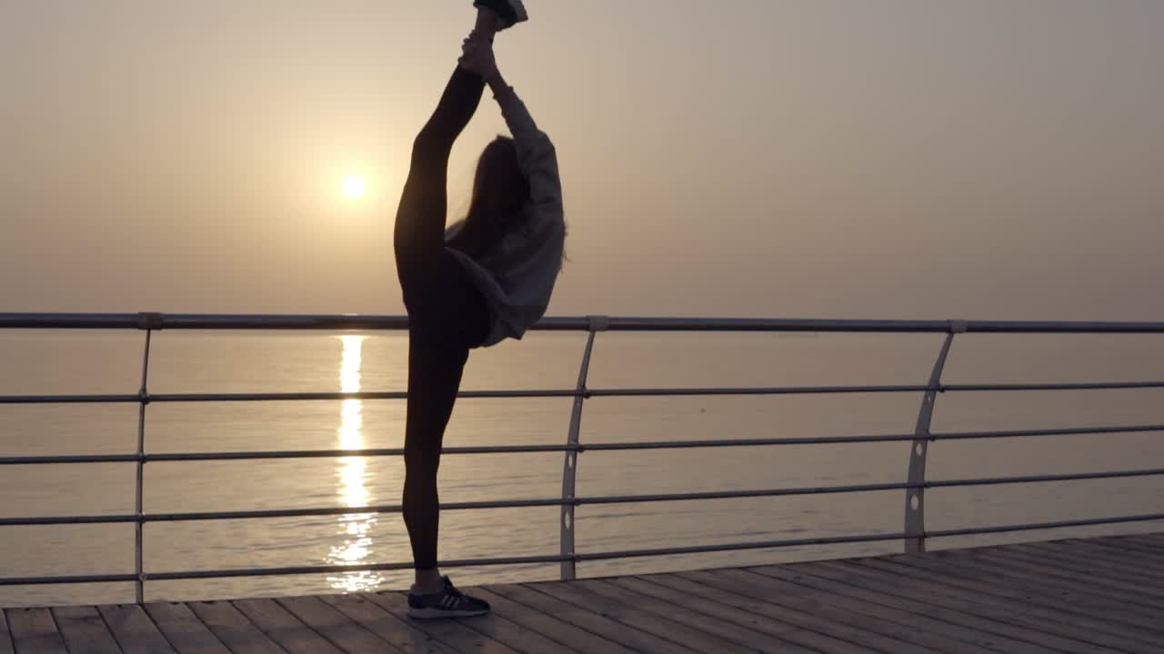 Long haired girl stretches her leg with a view of the sea ocean. Dawn Sunrise. Sportswear, leggings, hoodies. Dusk