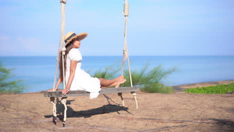 young woman in straw sunhat and white dress sitting and swinging on a wooden bench swing hanging on a tree at a tropical beach in thailand enjoying the sea view, slow motion