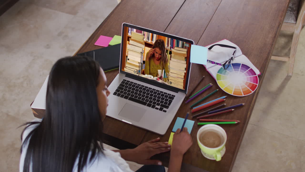 Profesora caucásica usando una laptop en videollamada con una estudiante, tomando apuntes.