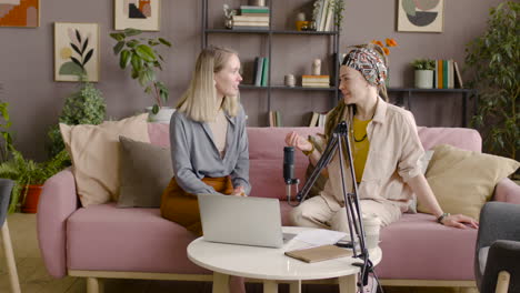 two women recording a podcast talking into a microphone sitting on sofa in front of table with laptop and documents 1