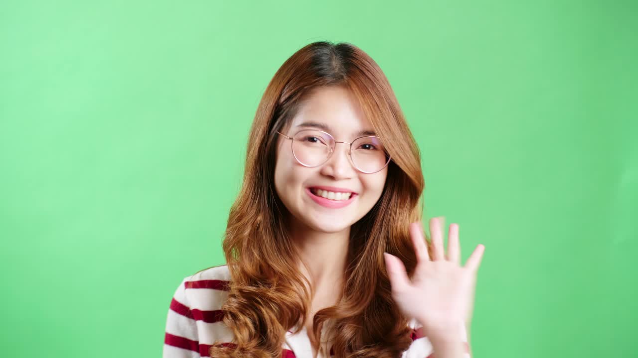 mujer asiática feliz con una camiseta a rayas con gafas mira la cámara y sonríe juguetonamente y saluda con un fondo de pantalla verde