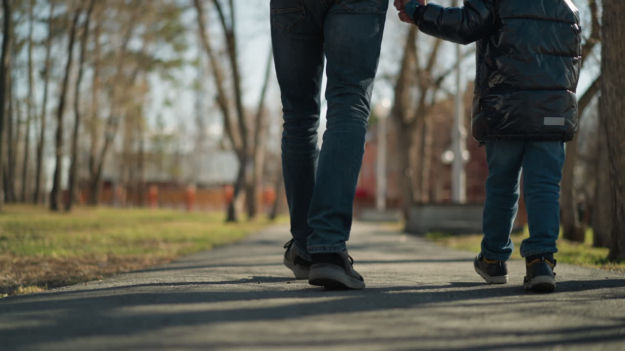 A close-up of father and son walking hand in hand on a pavement, the adult is wearing jeans and boots, while the child is dressed in a black leather jacket, jeans, and boots