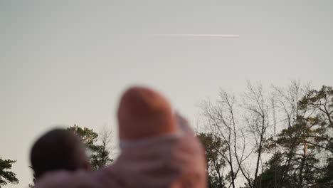 un hombre con una chaqueta verde lleva a su hija, vestida con una chaquita rosa y una gorra a juego, mientras ambos apuntan hacia un avión que atraviesa el cielo