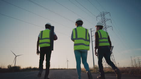 supervisores con cascos blancos y chalecos verdes caminando por un camino de grava bajo torres de transmisión junto a turbinas eólicas al atardecer