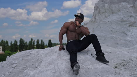 full length view of the athletic fit model with naked torso sitting at the sand. strong handsome man posing on blue sky background