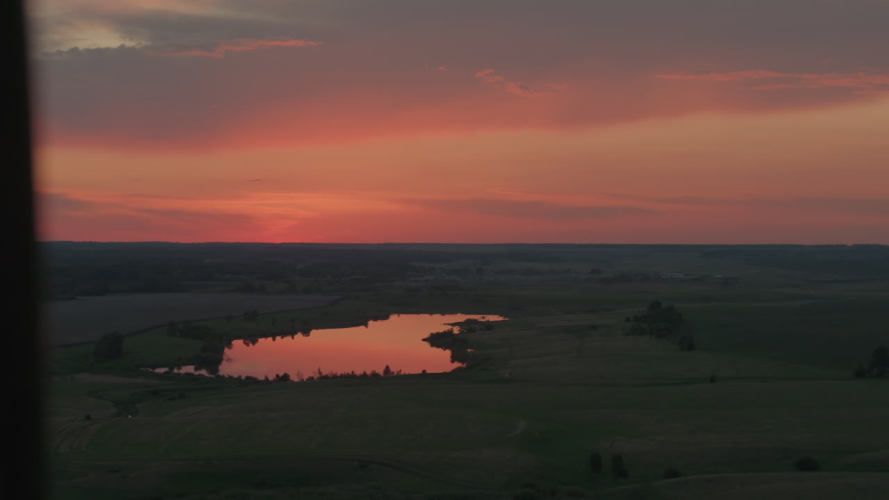 Scenic farmland panorama with tranquil lake mirroring vibrant sunset hues over vast fields while sun reflection shimmers on water surface beneath dramatic dusk clouds and winding creek