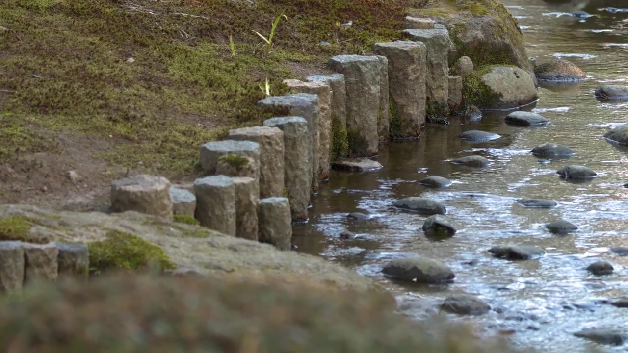 A peaceful stream flows over rocks past stone pillars in a tranquil Japanese garden.