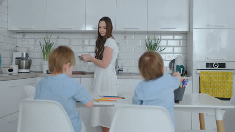 Two-boy-sitting-on-kitchen-light-draw-pencils-drawings-for-school-quests.-Mom-looks-at-the-children-and-smiles-in-the-kitchen