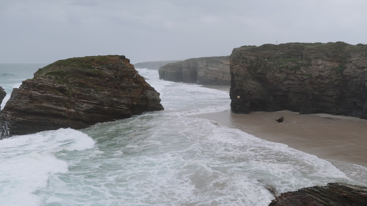 Ocean waves crashing on a sandy beach with large rock formations and cliffs under a cloudy sky