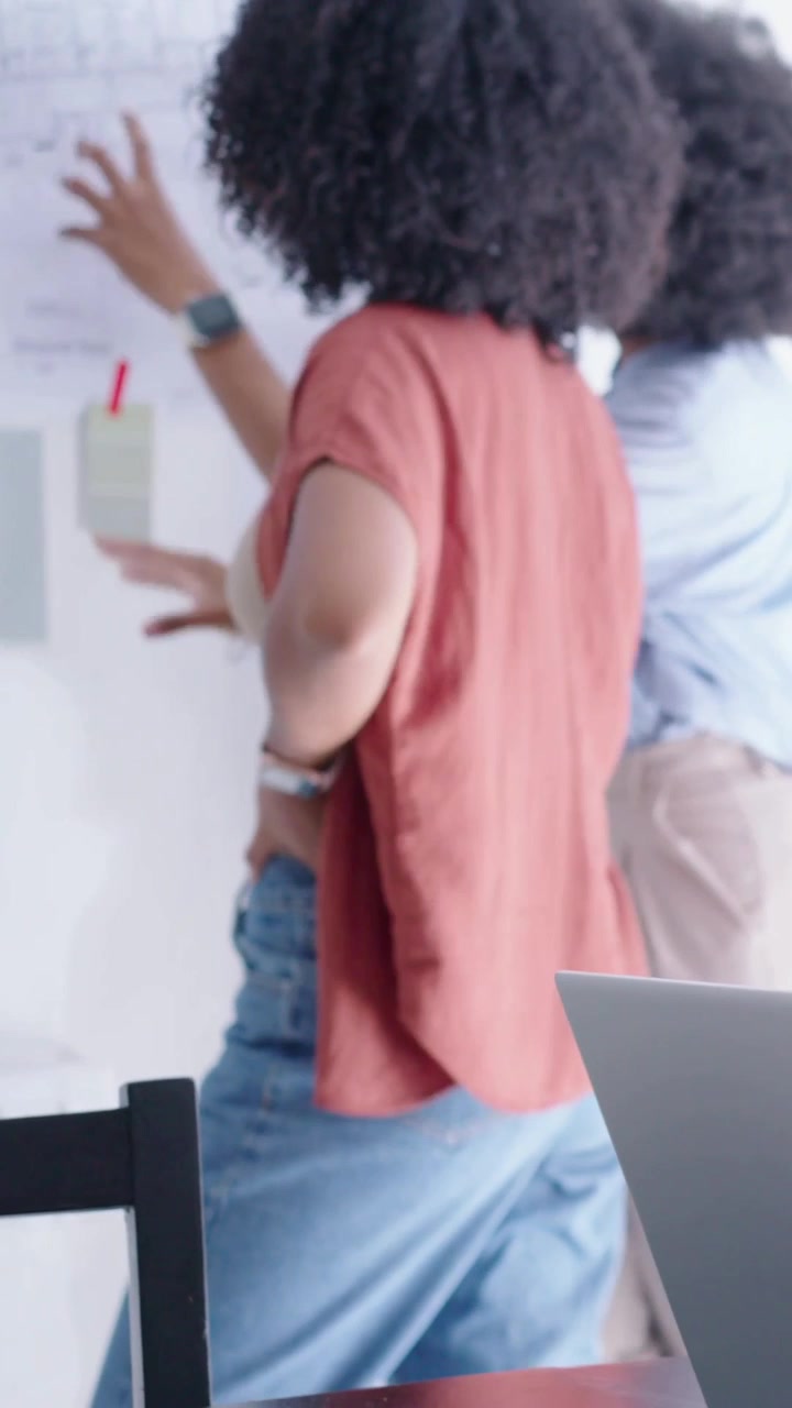 Vertical video: Team planning, woman in rust top pointing pinned notes at wall with red pen laptop