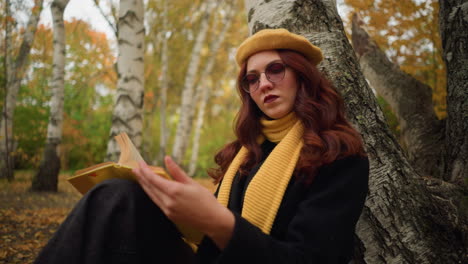 mujer pensativa con gafas de sol, boina amarilla y bufanda a juego revisa un libro mientras está sentada en un paisaje de otoño, rodeada de hojas y árboles, disfruta de la reflexión tranquila y la literatura