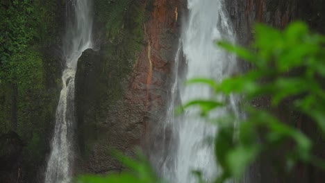 Twin-waterfall-falling-along-wet-rock-cliff-in-tropical-canyon,-bokeh-of-green-leaves