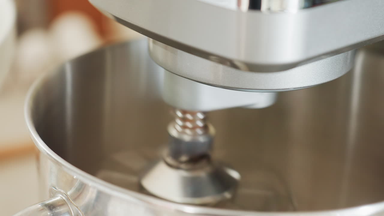 Close up of electric stand mixer in motion with spinning metal attachment inside shiny stainless steel mixing bowl, background softly blurred in bright modern kitchen