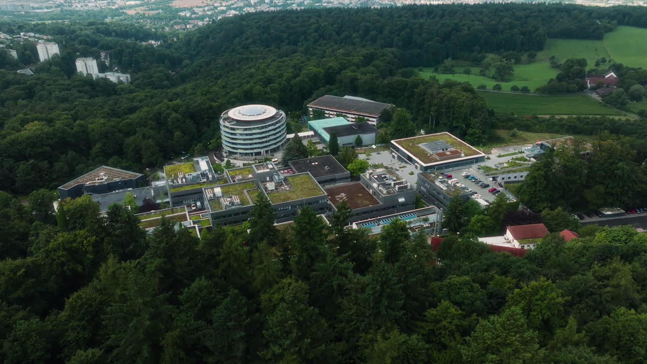 A drone performs a panning movement while keeping the DKFZ round building centered, then tilts up to reveal the forested hillside and the valley beyond