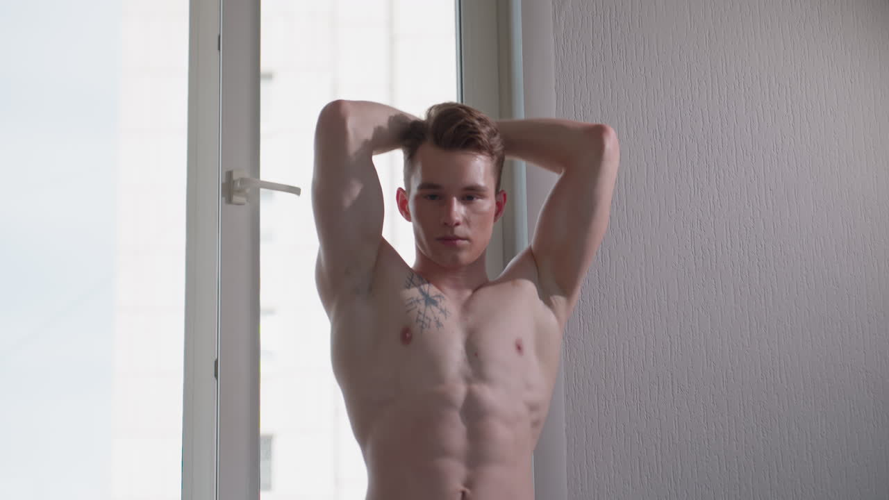 close up young man standing shirtless near window pulling resistant rope from behind, muscles flexed with intense focus, exercising with determination and resilience in softly lit indoor environment