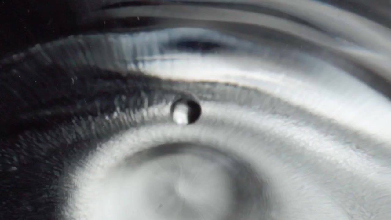 Primo piano macro di una goccia d'acqua che cade su una superficie liquida creando un motivo a increspature circolari al rallentatore. Movimento fluido astratto che mostra onde lisce e una texture riflettente.