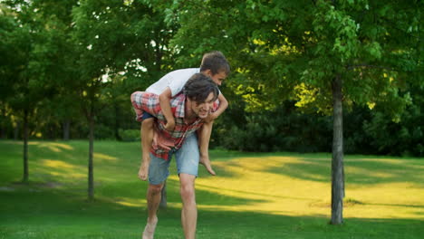son jumping on father back in green park. joyful man giving boy piggyback riding