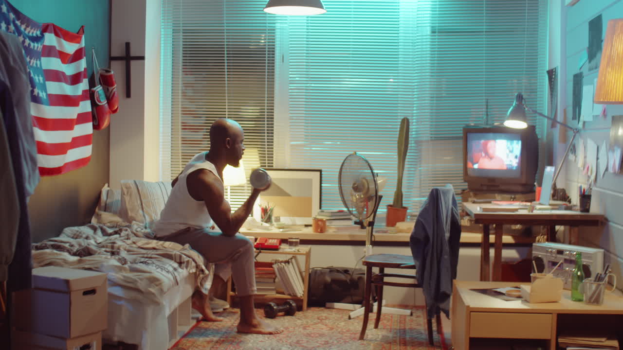 아프리카계 미국인 남자가 tv를 보고 집에서 덤벨 컬을 하고 있습니다.