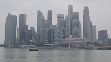 Plano-General:-La-Cámara-Estática-Captura-La-Impresionante-Arquitectura-Del-Horizonte-De-Singapur-En-Un-Día-Gris,-Con-Agua-Y-Barcos-En-Primer-Plano