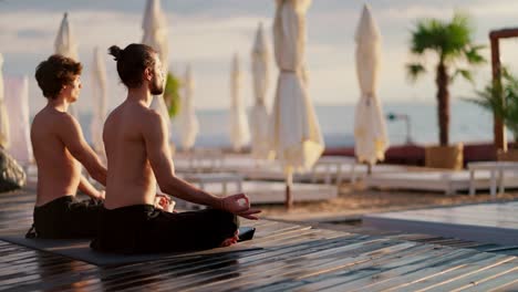 Two-bare-chested-guys-in-black-shorts-sit-and-meditate-on-a-private-beach-in-the-morning.-Harmony-of-body-and-mind