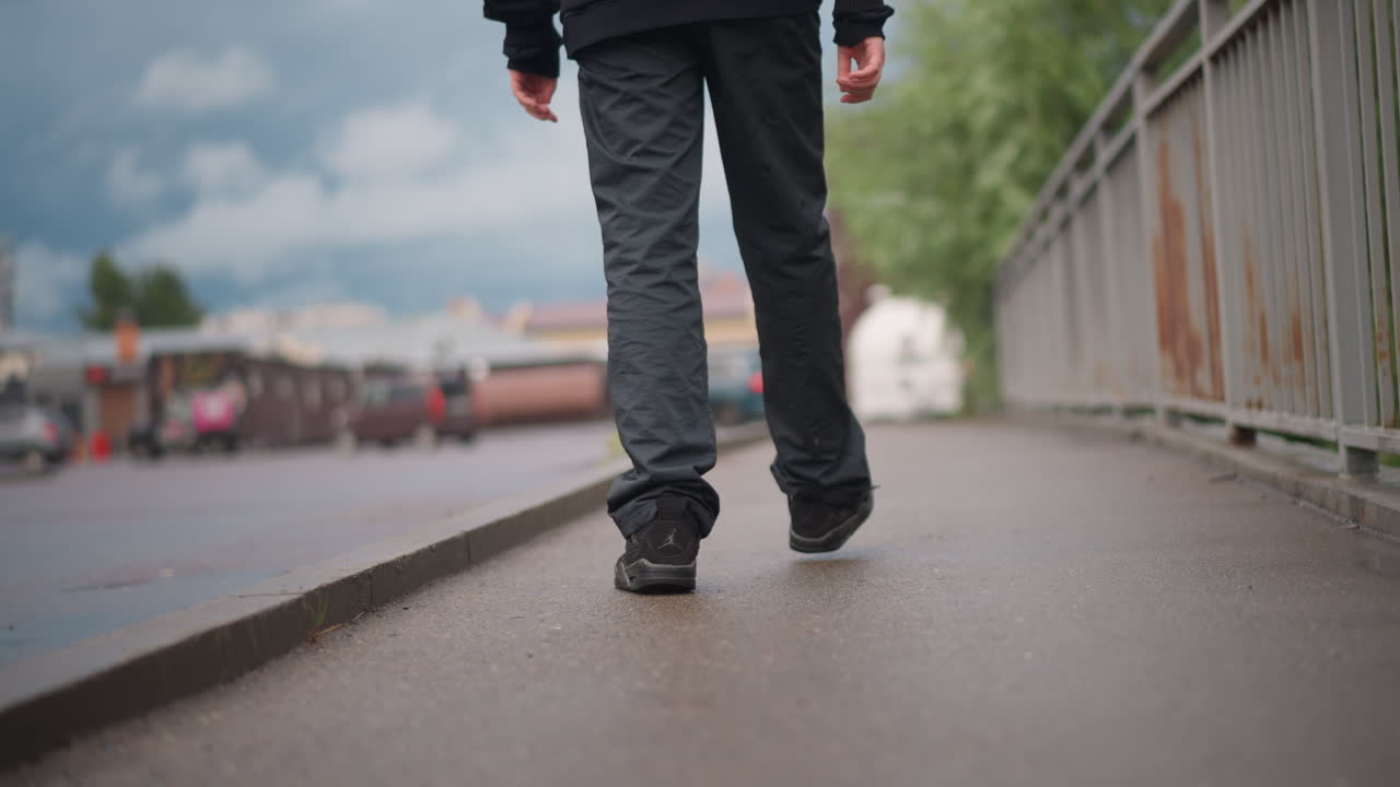 Close-up de sapato fazendo um pequeno respingo em concreto molhado pela chuva, cadarço desamarrado e sola texturizada em foco, vista cinematográfica em ângulo baixo de caminhante solitário em ponte com fundo nublado e poças refletoras