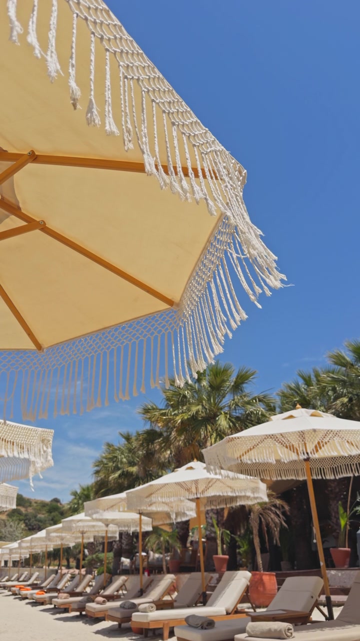 Splendida ripresa 4K dal basso di ombrelloni in macramè bianchi con frange in un resort di lusso sulla spiaggia. Presenta una fila organizzata di lettini, palme tropicali e un cielo azzurro limpido