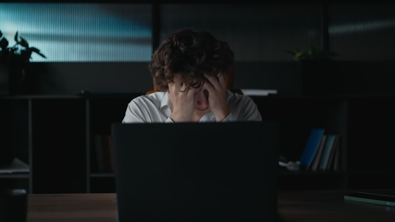 A guy with curly hair in a white shirt covers his face with his hands and shakes his head to the sides during a difficult task at work in the office