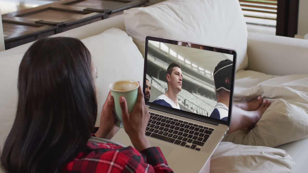 Composite of woman sitting at home holding coffee watching rugby match on laptop