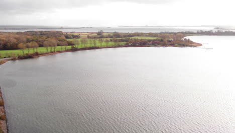 Cars-driving-on-road-between-Schlei-sea-and-tree-line-in-autumn,-tilt-up-aerial