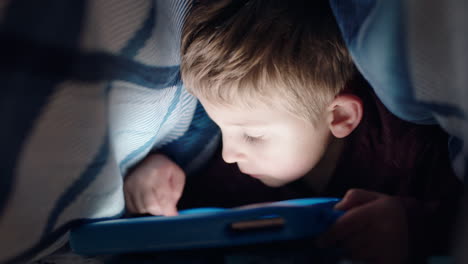 Un-Niño-Feliz-Usando-Una-Tableta-Digital-Bajo-Una-Manta-Disfrutando-Aprendiendo-Con-Tecnología-De-Pantalla-Táctil-Jugando-Juegos-Divirtiéndose-A-La-Hora-De-Dormir