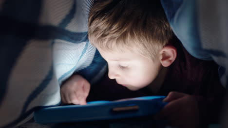 happy little boy using digital tablet computer under blanket enjoying learning on touchscreen technology playing games having fun at bedtime