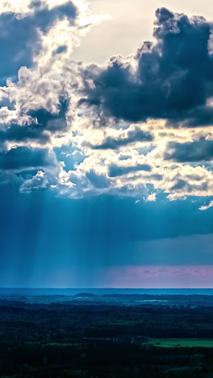 Il time-lapse verticale mostra nuvole soffici in rapido movimento che si spostano su un cielo blu intenso con scie di pioggia rosa su vasti campi e foreste rurali lettoni.