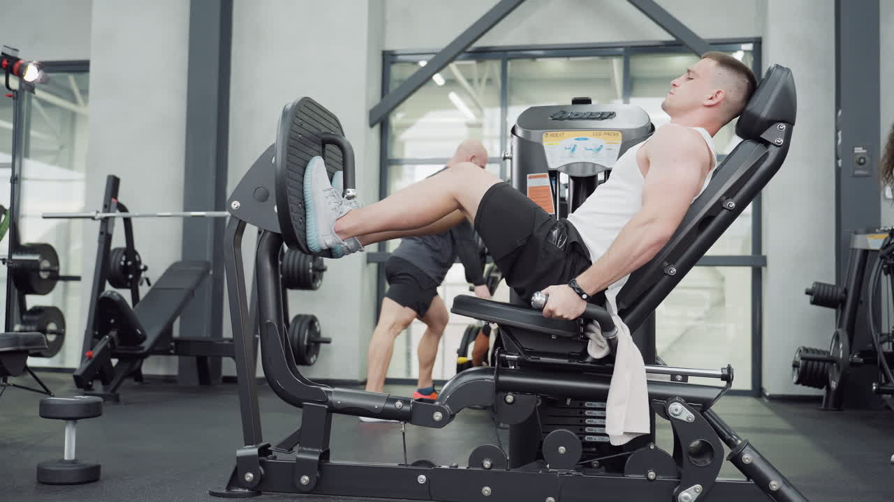 Gym junkie working out on leg press machine while instructor exercises in background under bright modern gym lighting showing focused strength, powerful form and effort during lower body workout