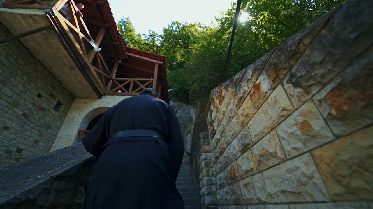 Following the priest wearing black robe. Orthodox monk going up the stone stairs of the monastery. Low angle view.
