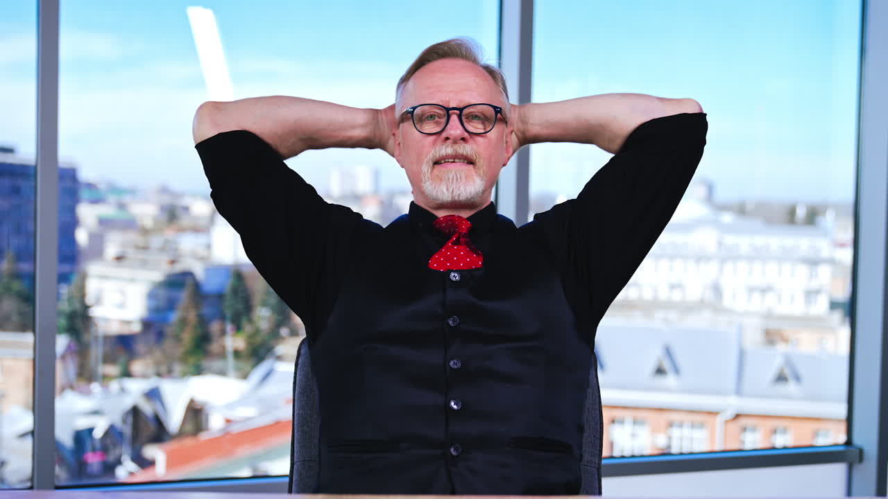 Man having a break from work in the office. Smiling positive businessman sits with his hands behind his head, takes off his eyeglasses and relaxes. Cityscape in window at backdrop in blur.
