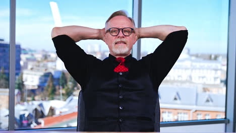 Man having a break from work in the office. Smiling positive businessman sits with his hands behind his head, takes off his eyeglasses and relaxes. Cityscape in window at backdrop in blur.
