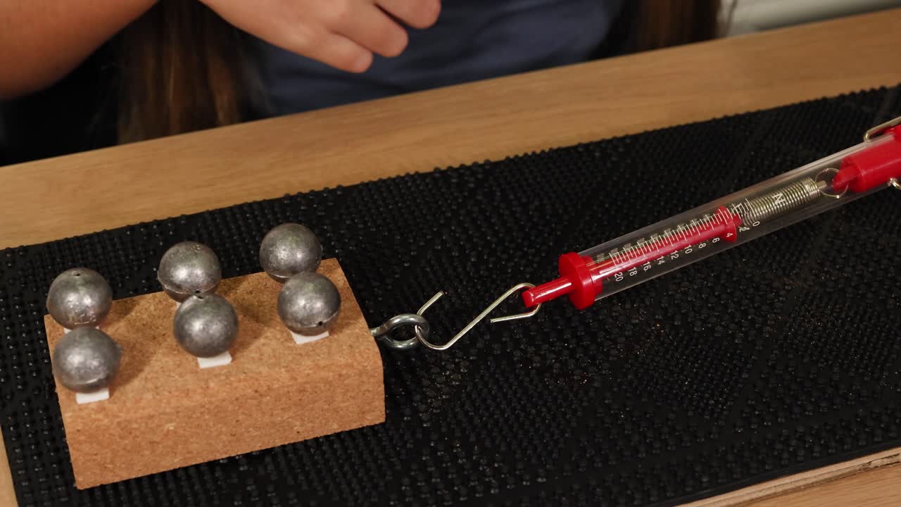 A hand uses a spring scale to measure force needed to drag weights on a cardboard surface, illustrating friction principles