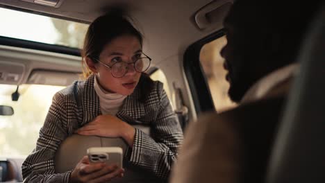 A-confident-brunette-girl-in-round-glasses-in-a-gray-jacket-communicates-with-a-man-with-Black-skin-in-a-brown-jacket-during-her-business-meeting-and-business-trip-in-a-modern-car-interior